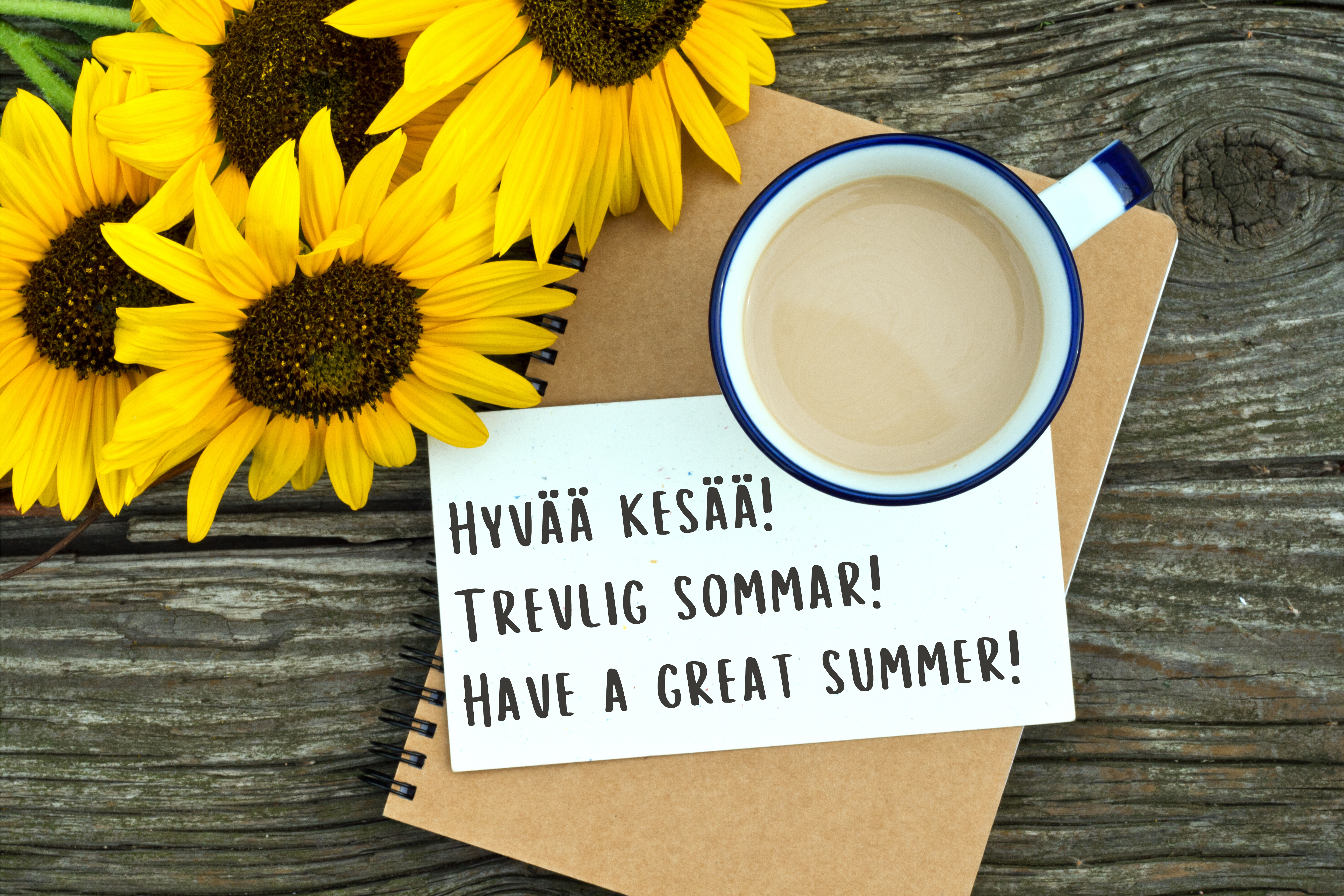 Pöydällä auringonkukkia, kahvikuppi ja lappu jossa lukee "Hyvää kesää! Trevlig sommar! Have a great summer!"